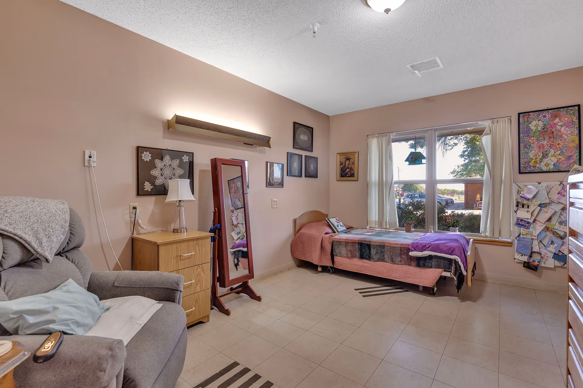 Private resident bedroom with a single bed, recliner, dresser, standing mirror and a large window overlooking the outside.