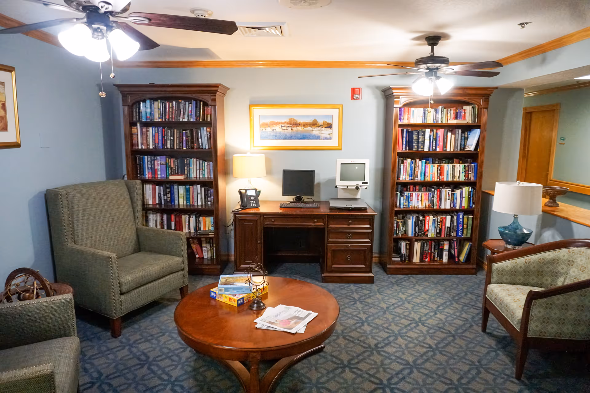 Cozy interior room with two tall wooden bookshelves filled with books, a wooden desk with a computer monitor and an old CRT monitor, a table lamp, and a telephone. The room has two ceiling fans with lights, a round wooden coffee table with newspapers and board games, and several upholstered chairs arranged around the table. The walls are painted light blue with wooden trim, and a framed picture hangs above the desk.
