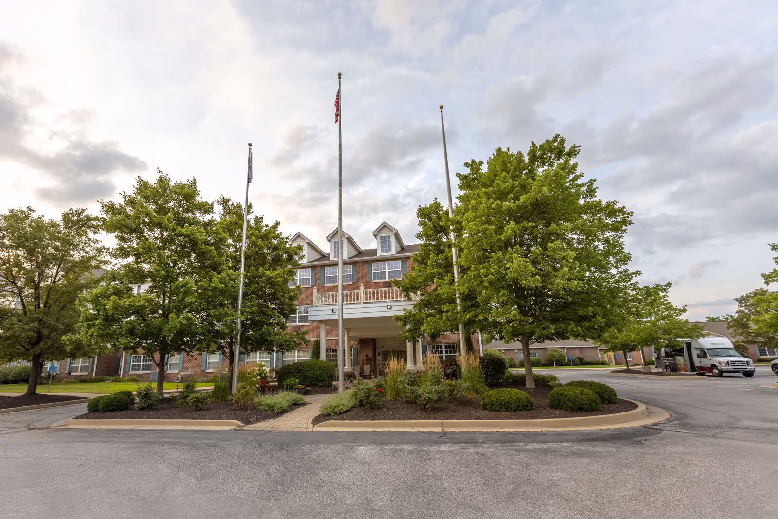 Front exterior view of a multi-story brick building with a covered entrance, surrounded by green trees and landscaping. Three flagpoles stand in front of the building, one with an American flag. A parking area and a white shuttle van are visible to the right.