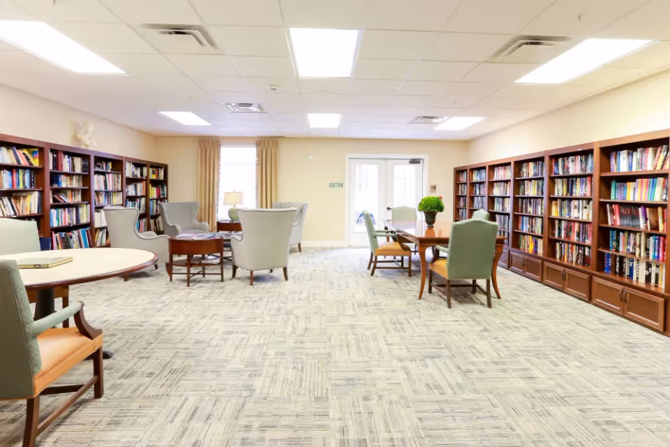 A spacious, well-lit library room with multiple bookshelves filled with books along the walls. The room features several comfortable armchairs arranged around small tables, and a larger table with chairs in the center. There is a window with beige curtains and a door with glass panels at the back of the room.