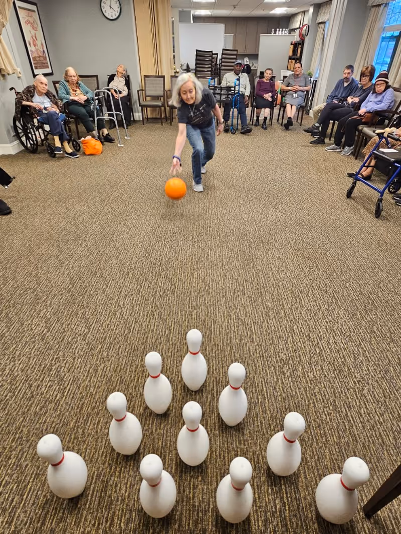 A group of elderly people seated in chairs arranged in a semicircle in a room with carpeted floor. One elderly woman is rolling an orange bowling ball towards a set of white bowling pins arranged on the floor. The room has a clock on the wall and some stacked chairs in the background.