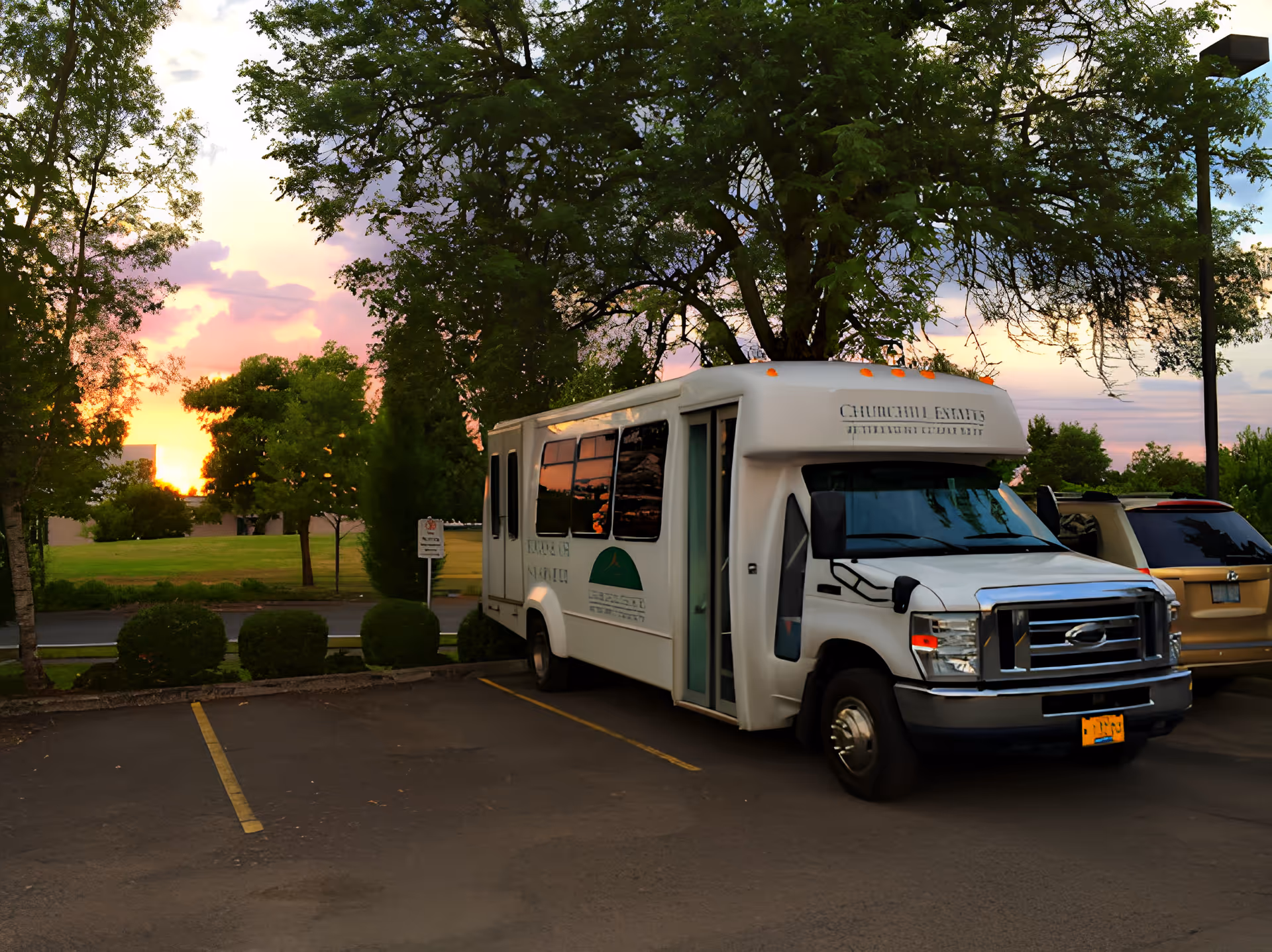 A white Churchill Estates retirement community shuttle bus parked in a lot at sunset with trees and a grassy field in the background.