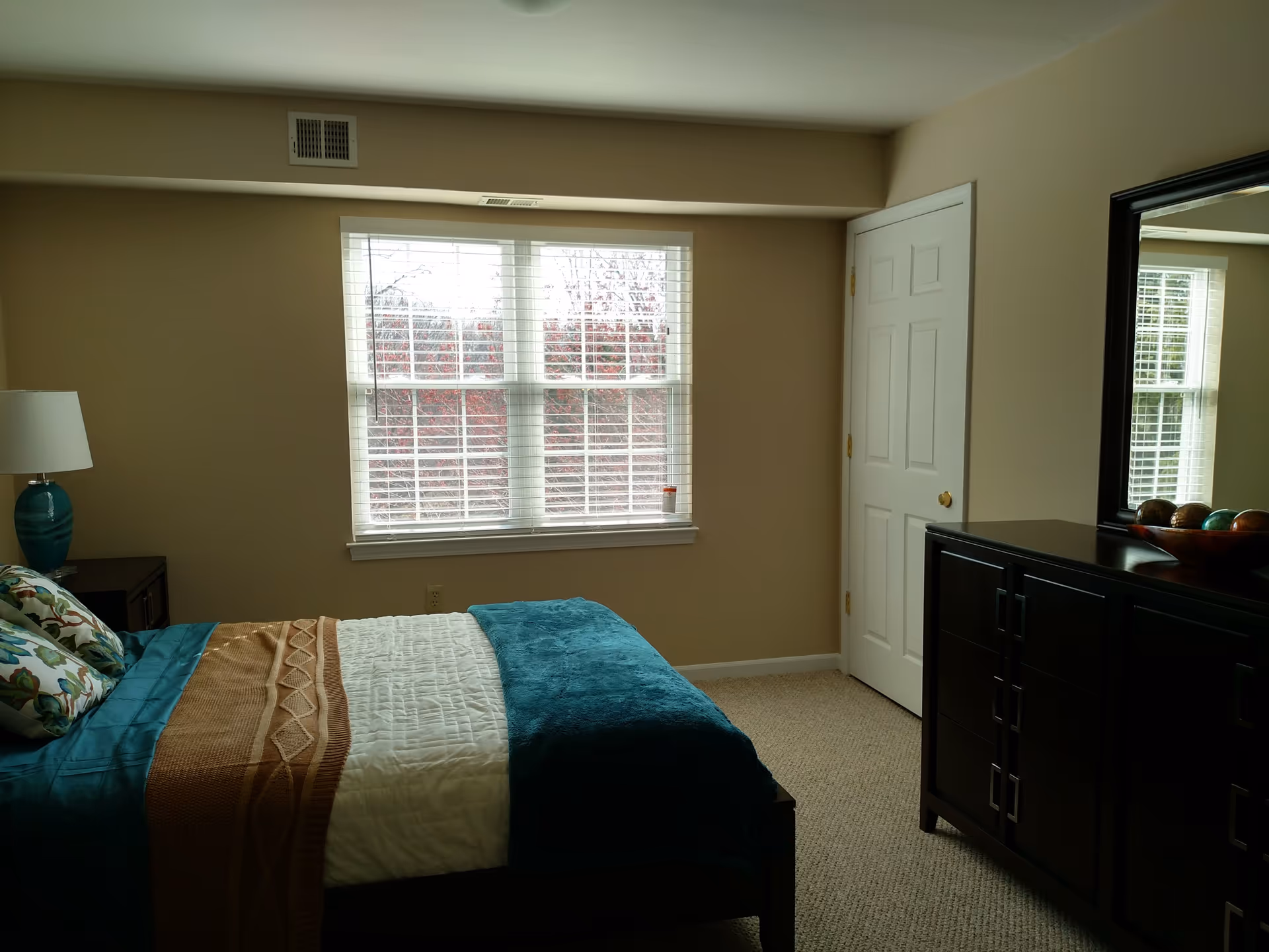 A bedroom with a bed covered in teal, white, and brown bedding, a nightstand with a teal lamp, a window with white blinds, a closed white door, and a dark wooden dresser with a large mirror and decorative bowl on top.