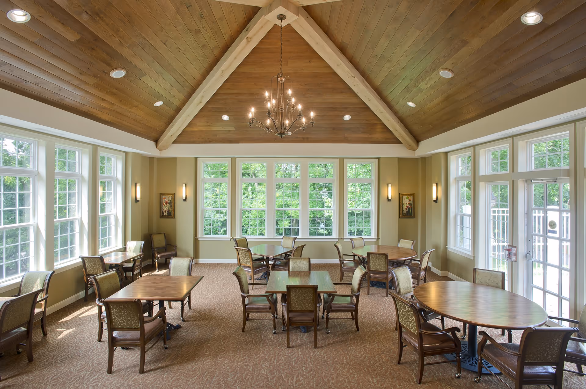 Sunlit communal dining room with multiple tables and chairs, large windows, and a wood-beamed vaulted ceiling with a chandelier.