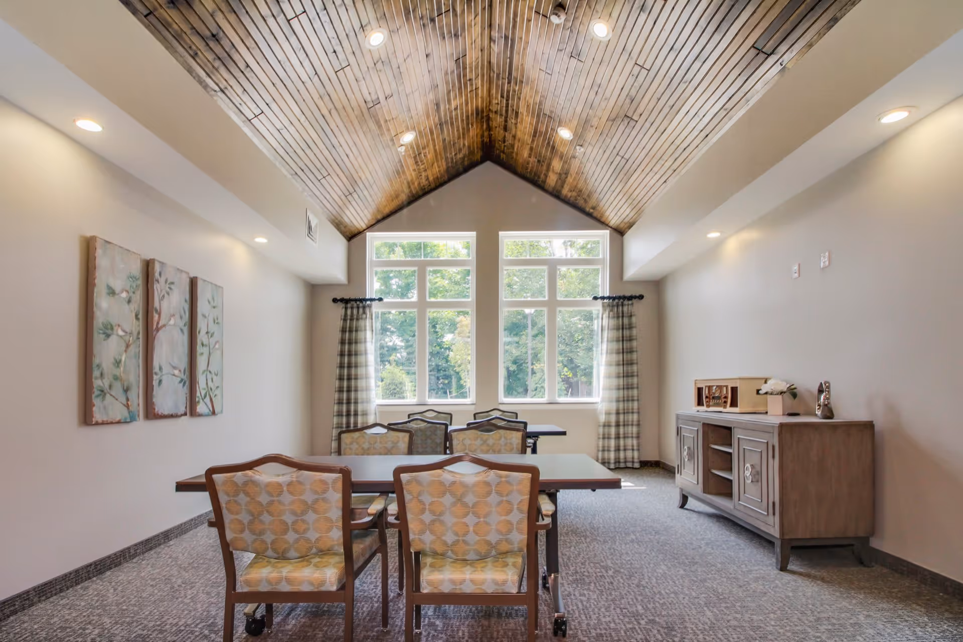 A bright room with a vaulted wooden ceiling and large windows letting in natural light. The room contains two tables with six patterned chairs arranged around them. On the right side, there is a wooden sideboard with decorative items on top. The walls are light-colored with three framed artworks on the left wall and plaid curtains on the windows.