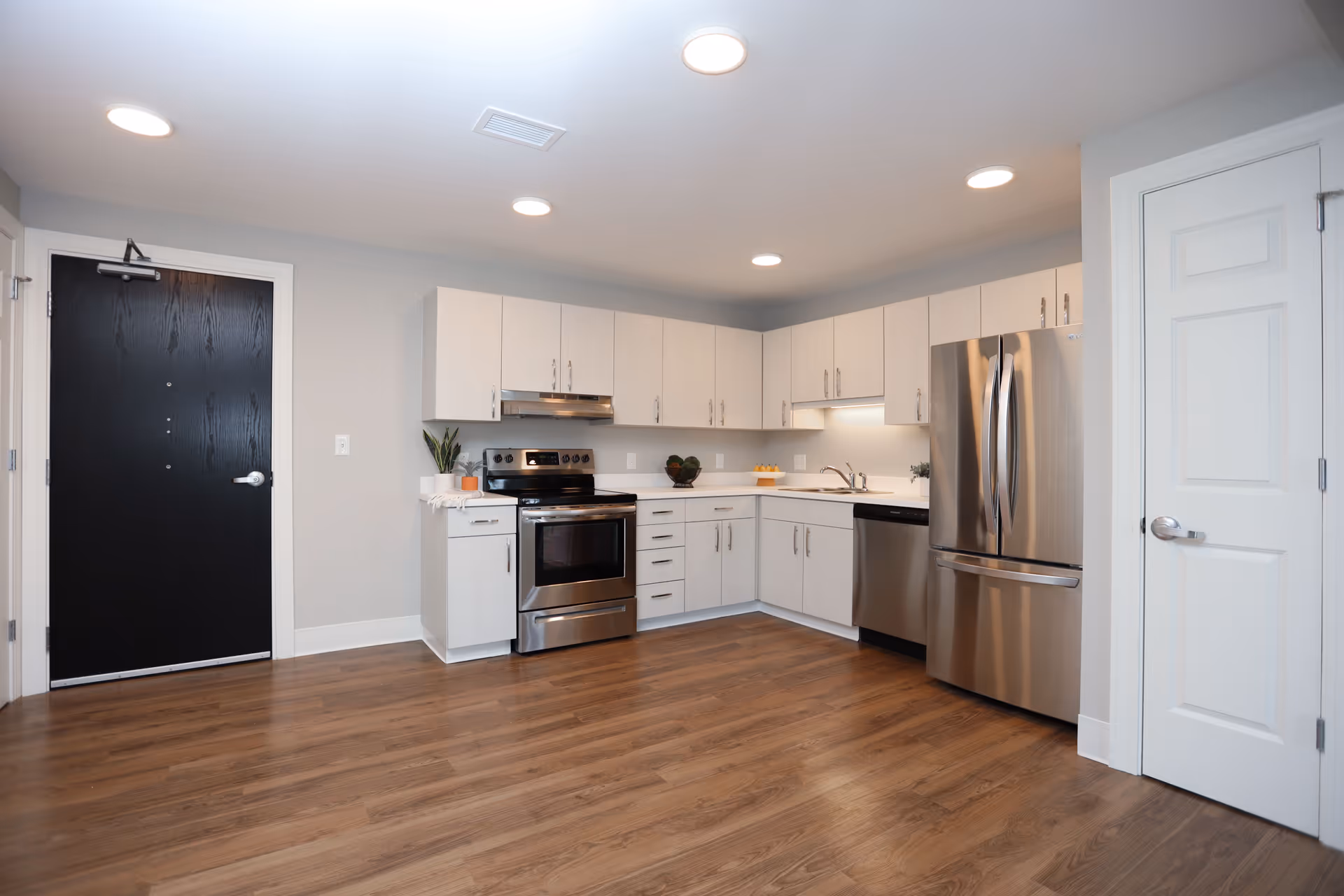 Modern kitchen with white cabinets, stainless steel appliances including a refrigerator, stove, and dishwasher, wood flooring, and recessed ceiling lights. There are two doors visible, one black and one white.