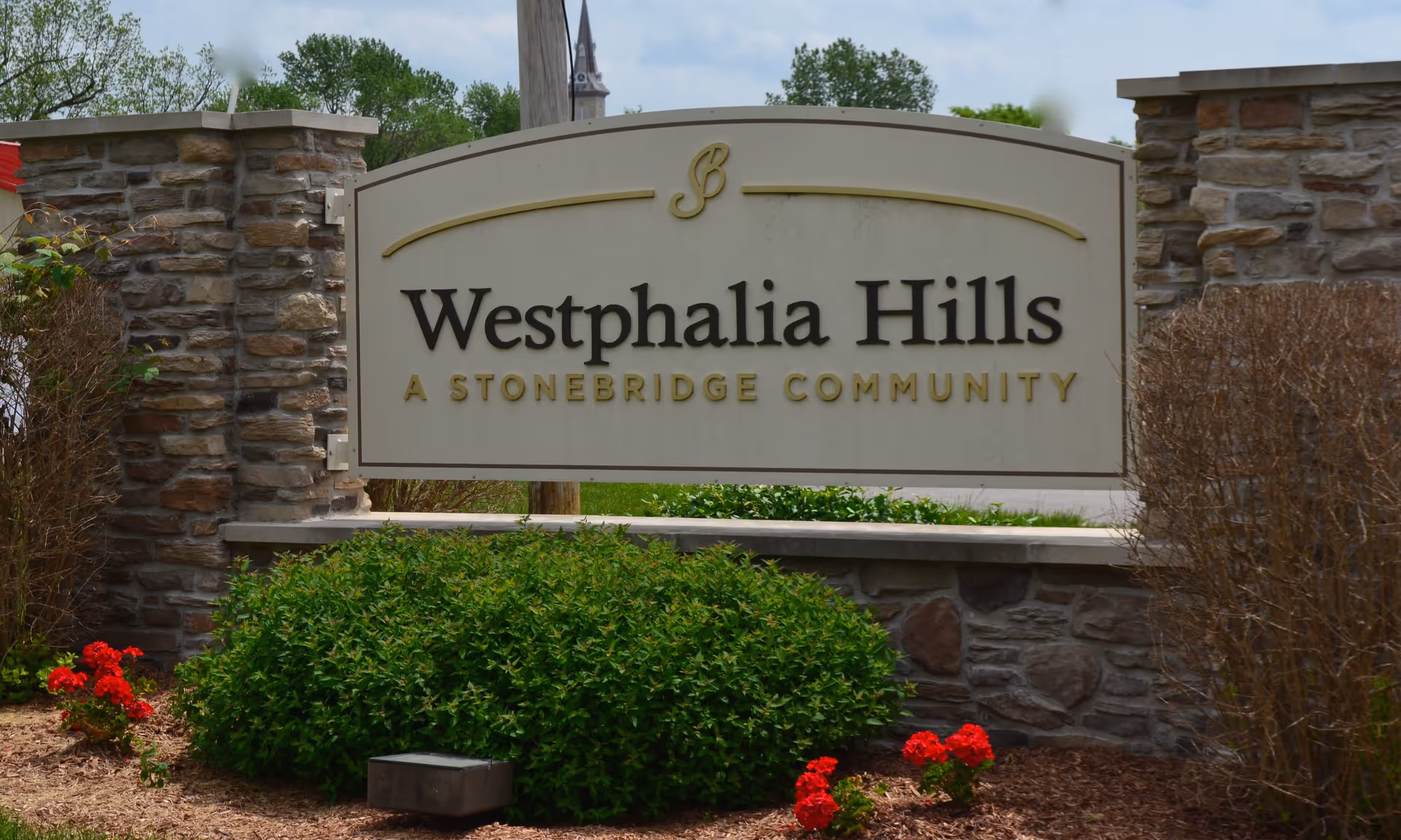 Stone sign for Westphalia Hills, a Stonebridge community, set between two stone pillars with green bushes and red flowers in front.