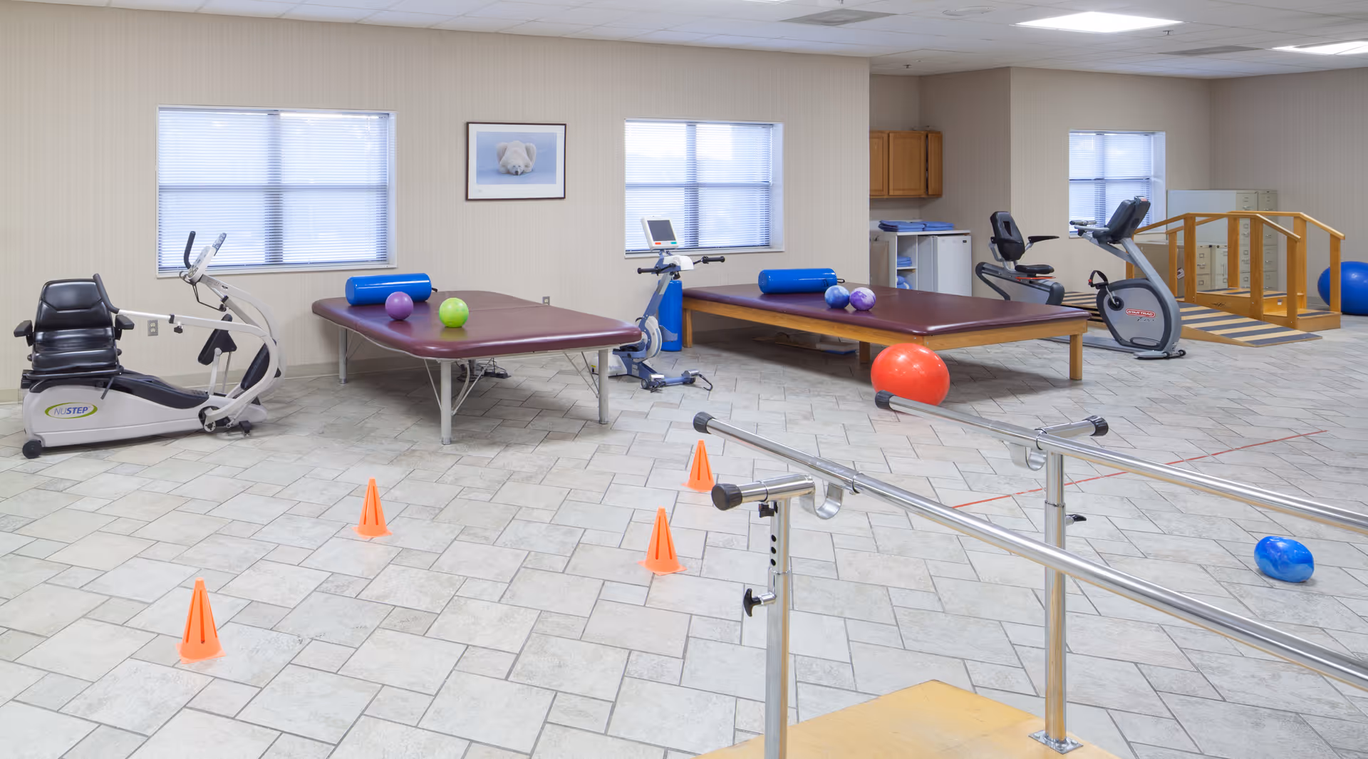 A physical therapy room with exercise equipment including stationary bikes, therapy tables with exercise balls, orange cones on the tiled floor, parallel bars for walking exercises, and a small wooden ramp. The room has beige walls, three windows with blinds, and a framed picture of a polar bear on the wall.