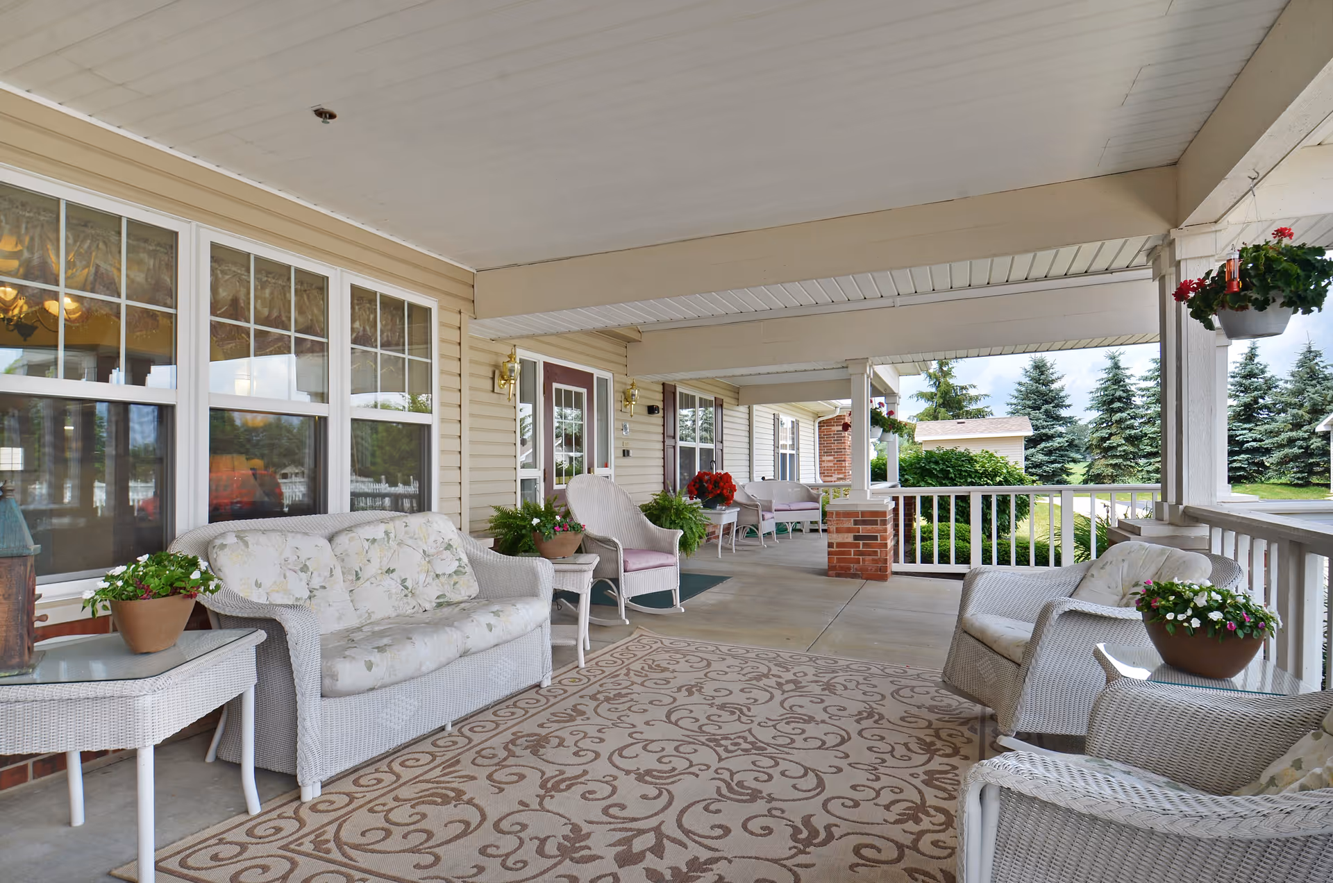 Covered outdoor porch area with white wicker furniture including a sofa, chairs, and side tables. The porch has a patterned rug, potted plants, hanging flower baskets, and overlooks a green lawn with trees in the background.