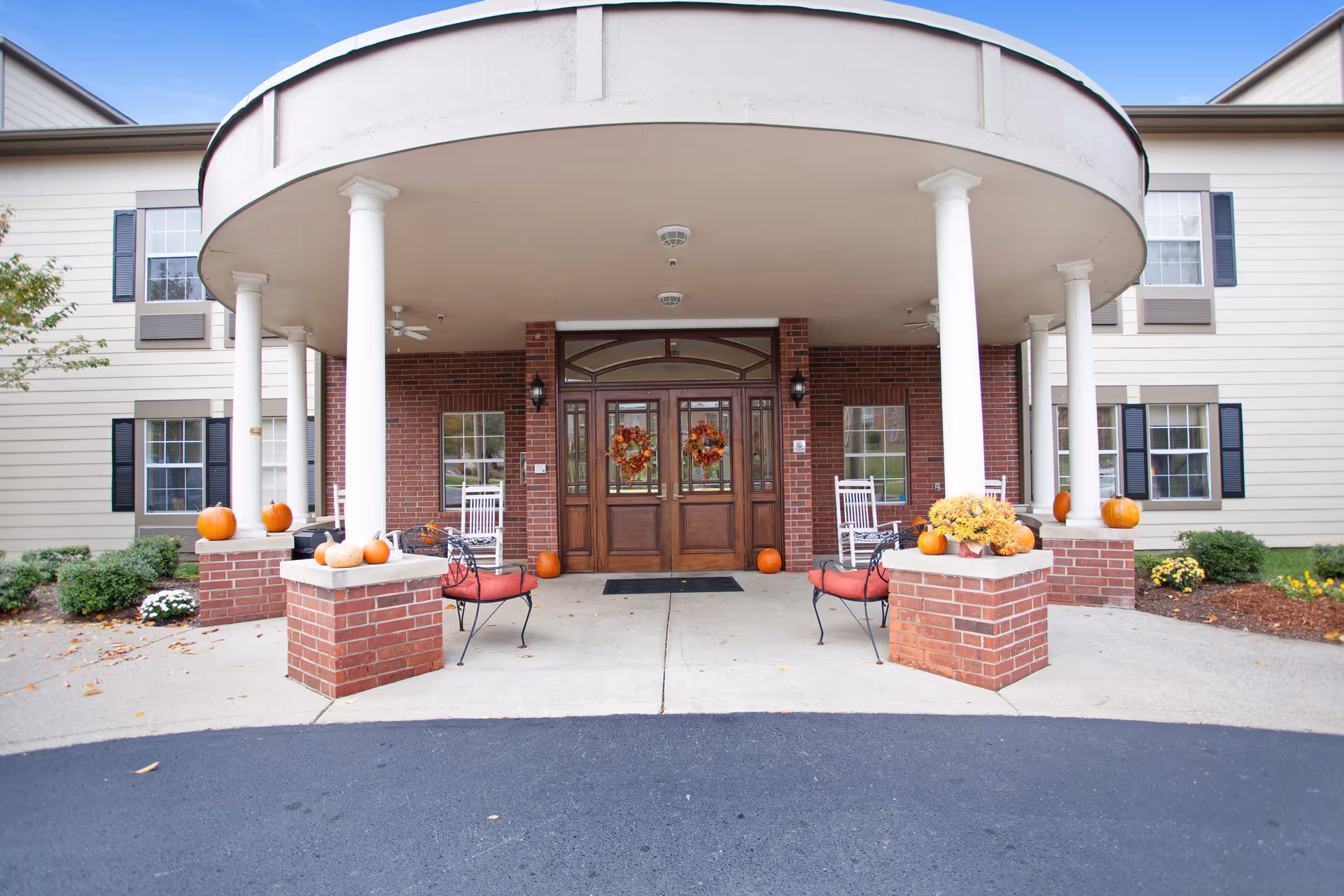 Front entrance of a senior living building with a covered circular portico, brick pillars, chairs, and autumn decorations including pumpkins.