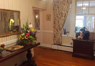 A cozy interior space with wooden flooring and beige wallpaper. A dark wooden table against the wall holds a large colorful flower arrangement and a plate of cookies. Above the table is a large mirror with a wooden frame. To the right, a floral curtain partially covers a window with white trim. A dark wooden cabinet with various items on top is positioned near the window.