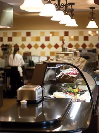 Interior view of a kitchen or food service area with a curved glass display case containing food items. Overhead lights hang above the counter, and a person in a white shirt and black apron is seen working in the background near a tiled wall with a diamond pattern.