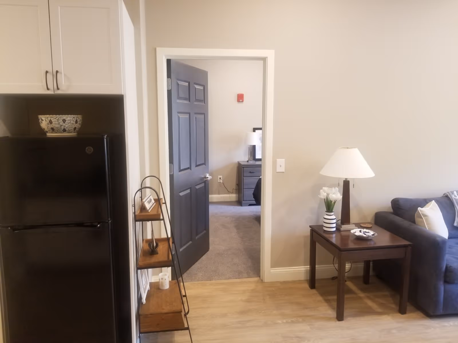 Interior view of a senior living facility showing part of a kitchen with a black refrigerator and white cabinets on the left, a small wooden shelf unit, an open door leading to a bedroom with a nightstand and lamp, and a living room area with a dark gray couch, a wooden side table with a lamp, a vase with white flowers, and a small dish.