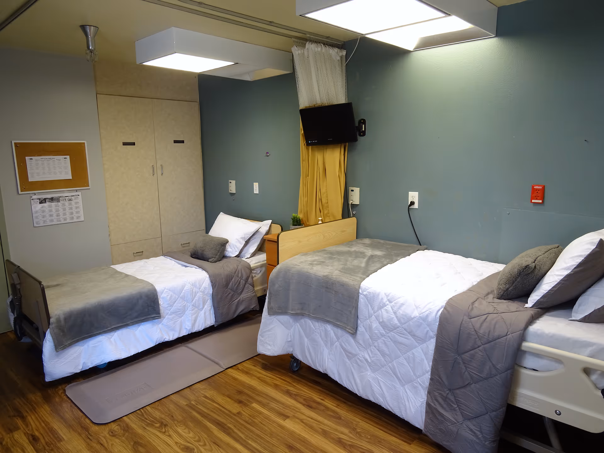 Two neatly made twin care beds in a shared senior facility room with wood flooring, wall-mounted TV and storage cabinets.