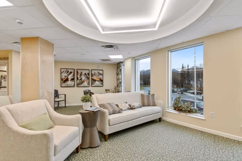 A bright and cozy senior living facility lounge area with beige walls and carpeted floor. The room features a beige sofa with multiple decorative pillows, a matching armchair with a green cushion, and a small round side table with a plant and a magazine. Large windows allow natural light to fill the space, and three framed pictures hang on the far wall. Additional chairs are visible in the background.