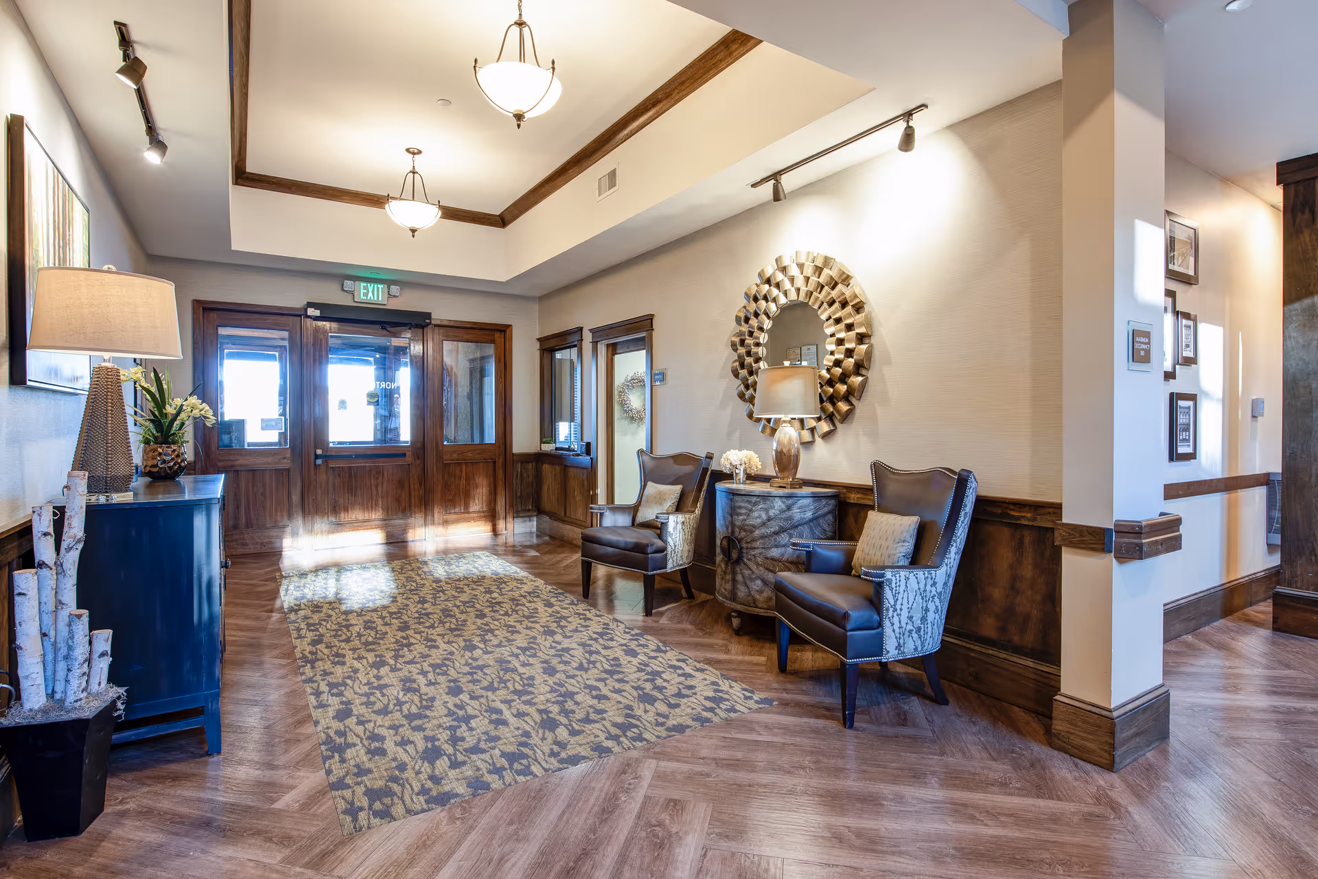 A bright and welcoming lobby area with wooden double doors at the entrance, a patterned rug on the wooden floor, two armchairs with cushions, a round decorative mirror on the wall, a small cabinet with a lamp and flowers, and additional furniture and decor along the walls.