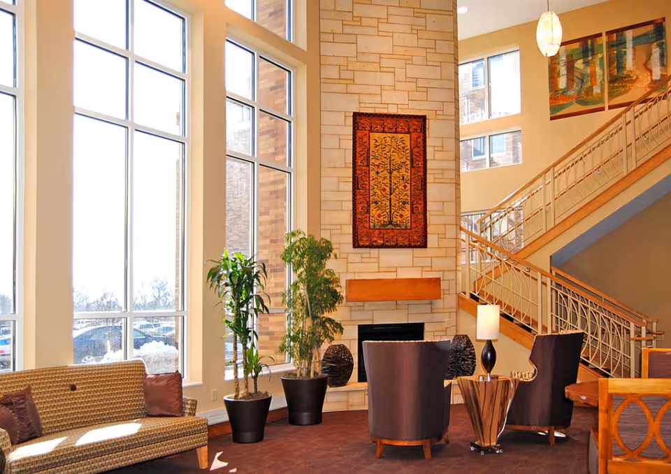A bright and spacious living room area with large floor-to-ceiling windows letting in natural light. The room features a patterned sofa with pillows, two dark armchairs around a small wooden table with a lamp, and two potted plants near a stone fireplace. Above the fireplace hangs a colorful tapestry. A staircase with a wooden handrail and decorative metal balusters is visible in the background.