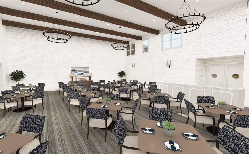 A spacious dining room with multiple square tables set with plates and napkins. Each table has four cushioned chairs with patterned backs. The room features white brick walls, large windows near the ceiling, wooden ceiling beams, and circular chandeliers. There are plants and decorative artwork on the far wall.