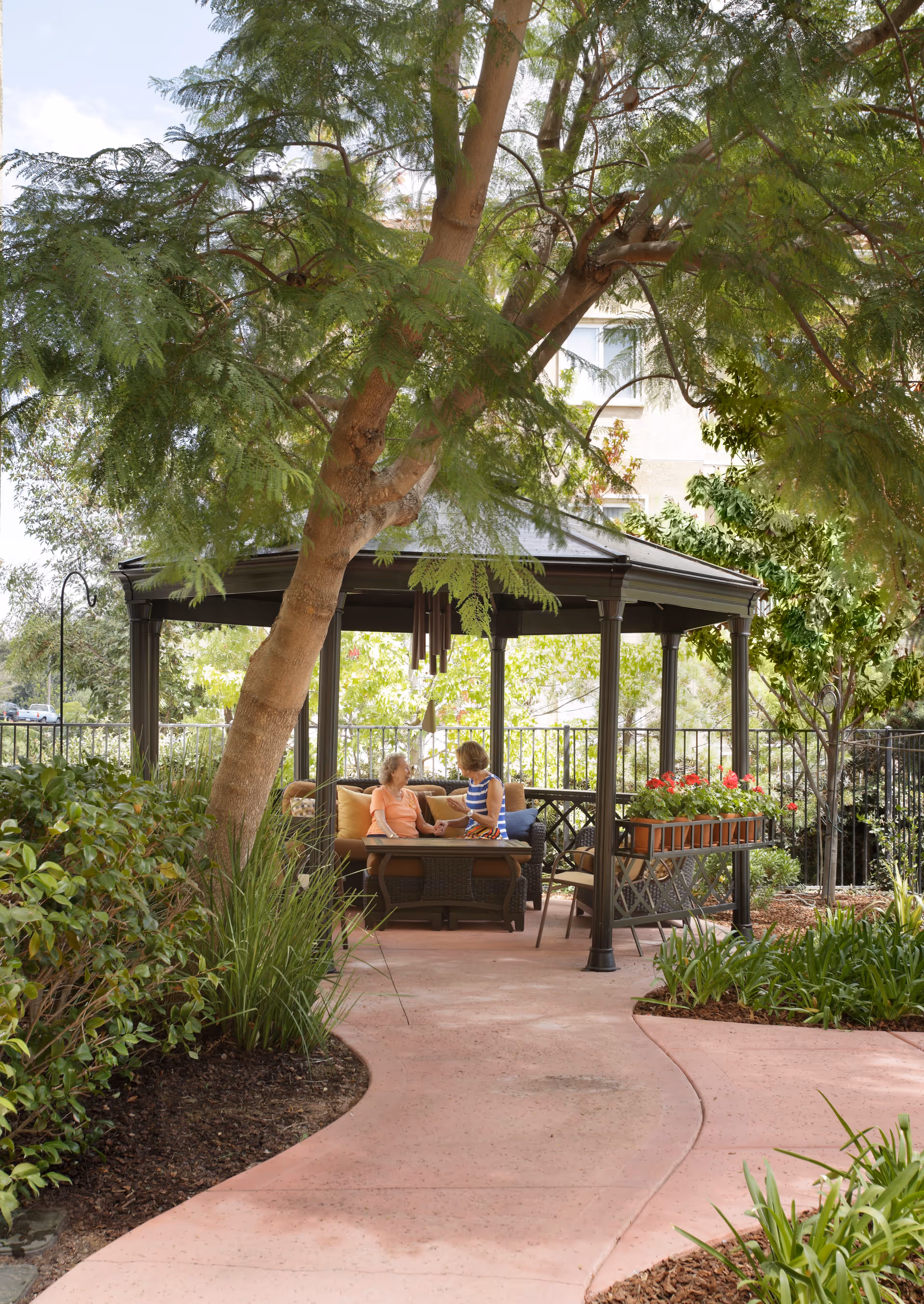 Two women sitting and talking under a black gazebo in a garden area with lush greenery and a curved pink concrete pathway leading to the gazebo.
