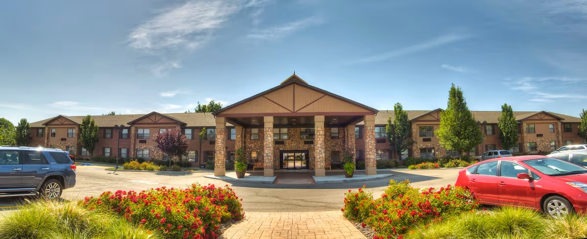 Front exterior view of Garden Plaza of Valley View, a two-story building with a covered entrance supported by stone pillars, surrounded by landscaped greenery and parked cars under a clear blue sky.
