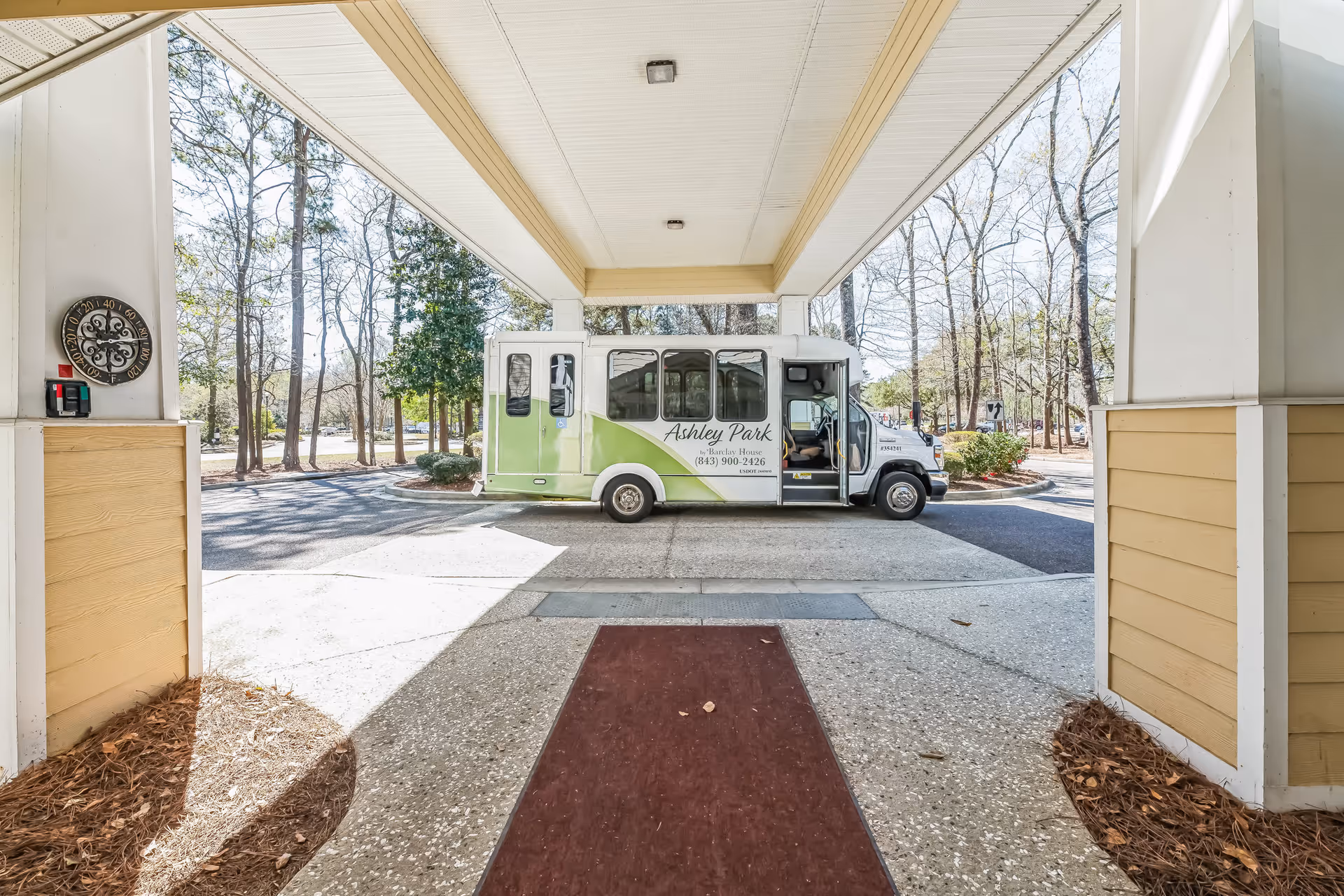 A green-and-white Ashley Park shuttle parked under a covered entrance/porte-cochere framed by columns and trees.