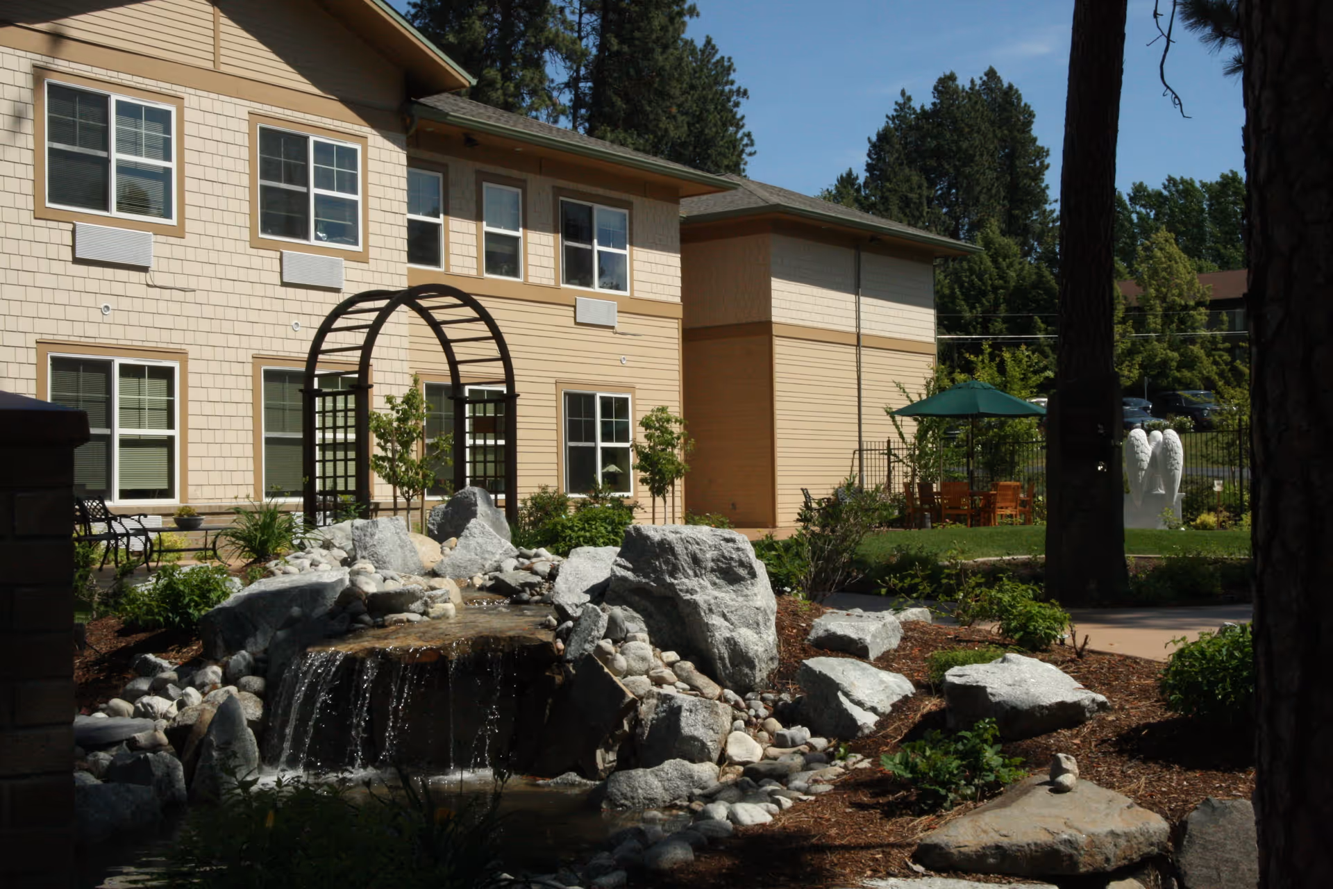 Outdoor garden area at Orchard Ridge Senior Living featuring a small waterfall flowing over rocks, a wooden arch trellis, benches, green plants, trees, and a patio with a table and umbrella. The building with multiple windows is visible in the background under a clear blue sky.
