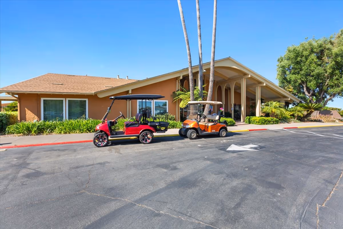 Exterior view of a single-story building with a sloped roof and large windows, surrounded by greenery and palm trees. Two golf carts, one red and one orange, are parked on the asphalt driveway in front of the building under a clear blue sky.