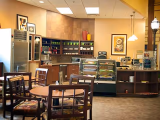 Small cafeteria-style dining area with tables and chairs facing a service counter with a refrigerated display case, coffee machines, and shelving.