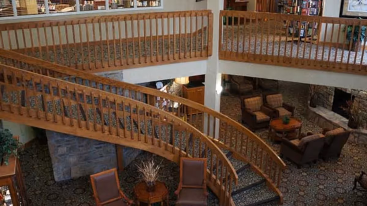 Interior view of a senior living facility featuring a curved wooden staircase with railings, a patterned carpet, and a seating area with armchairs and a small table near a stone fireplace.