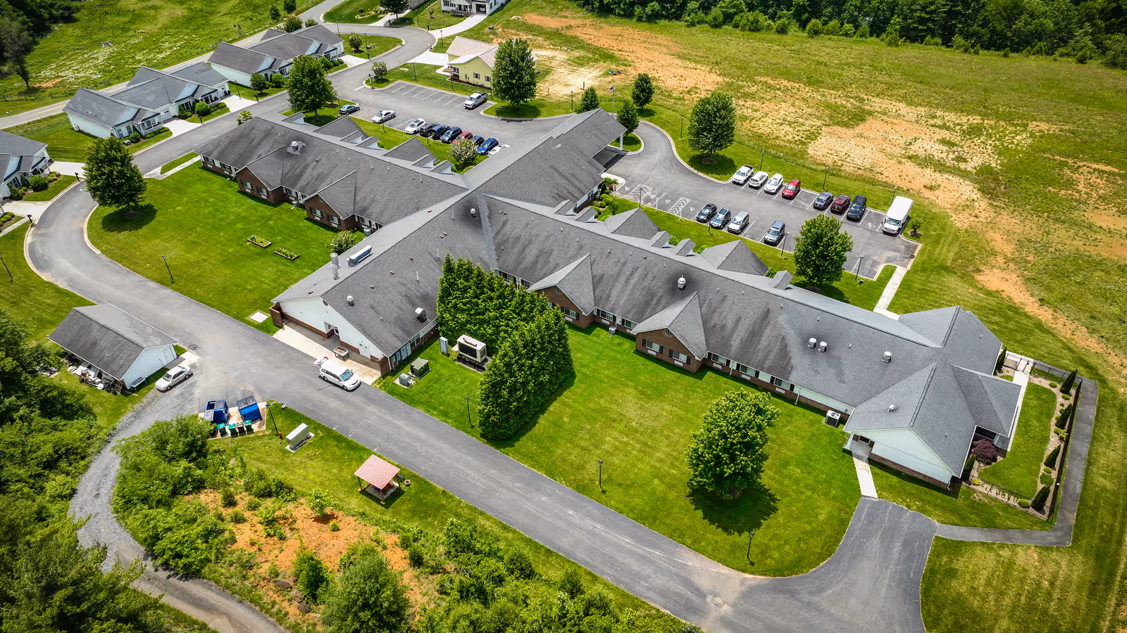 Aerial view of Forest Ridge Assisted Living facility showing a large single-story building with multiple wings surrounded by green lawns, trees, parking lots with cars, and a curved driveway. The facility is situated in a semi-rural area with open fields nearby.