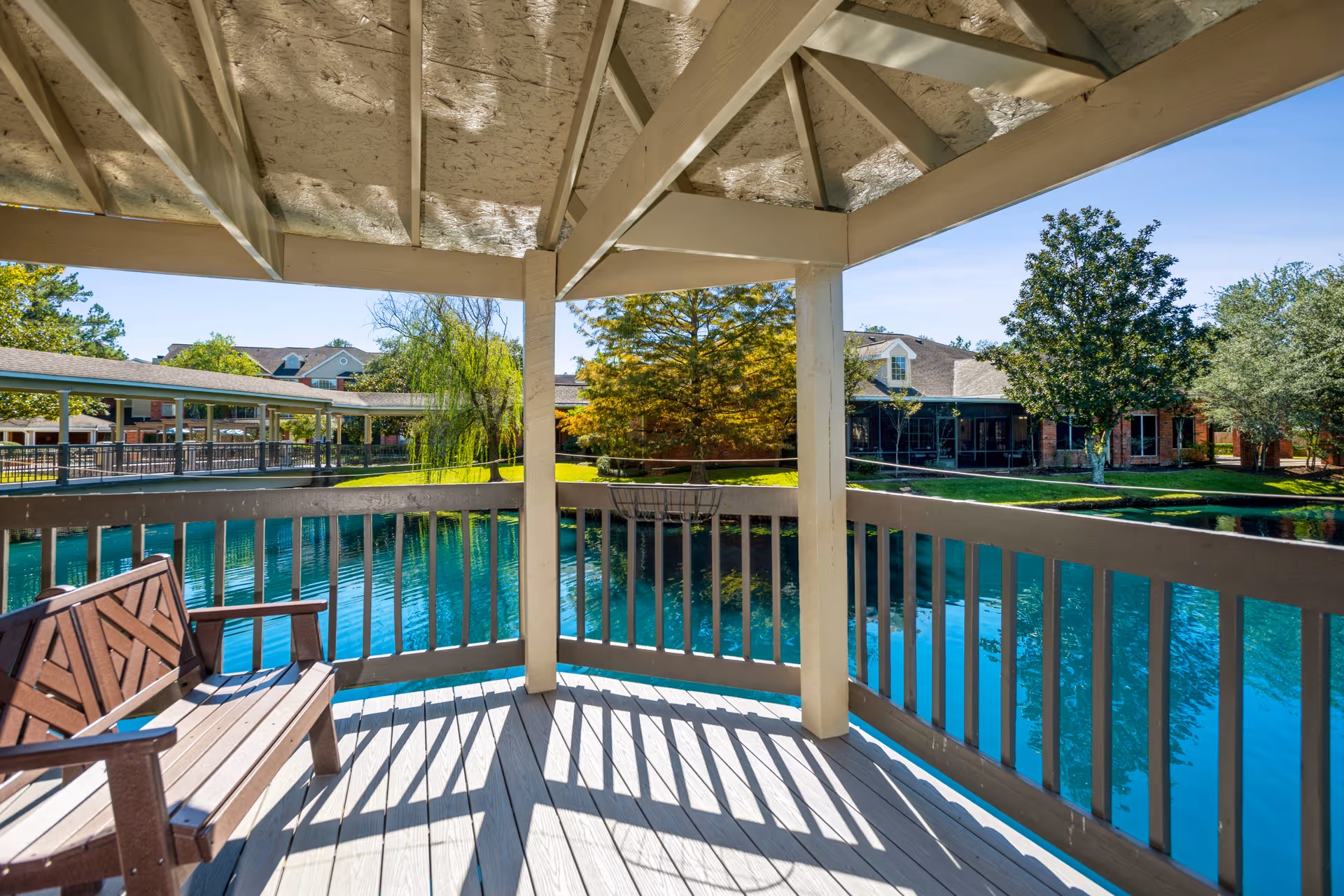 View from a covered wooden gazebo overlooking a serene pond with clear blue water, surrounded by green trees and buildings in the background under a clear blue sky.