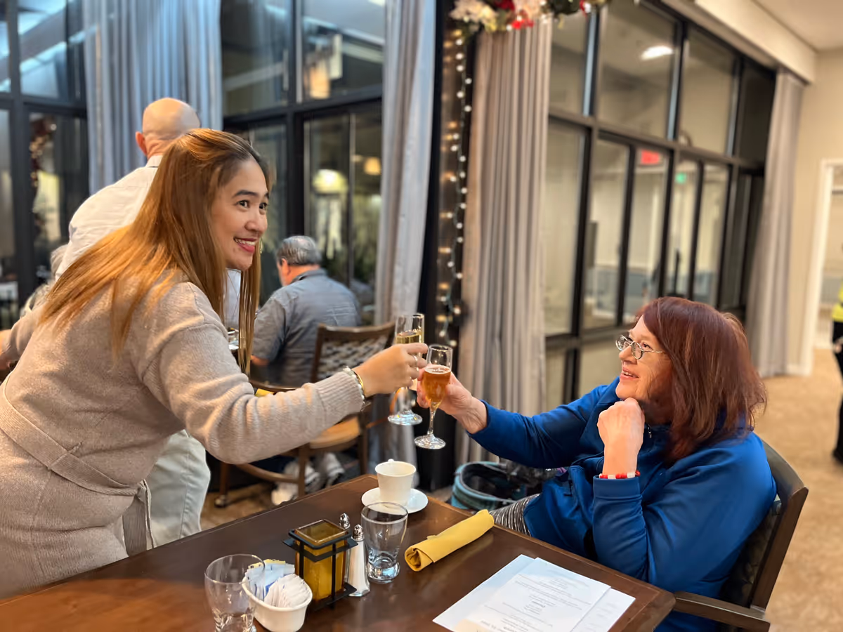 Two women smiling and clinking glasses in a dining area. One woman with long light brown hair is standing and leaning forward, while the other woman with red hair and glasses is seated at a table with a menu, napkin, and glassware. Other people are visible in the background, and large windows with curtains are behind them.