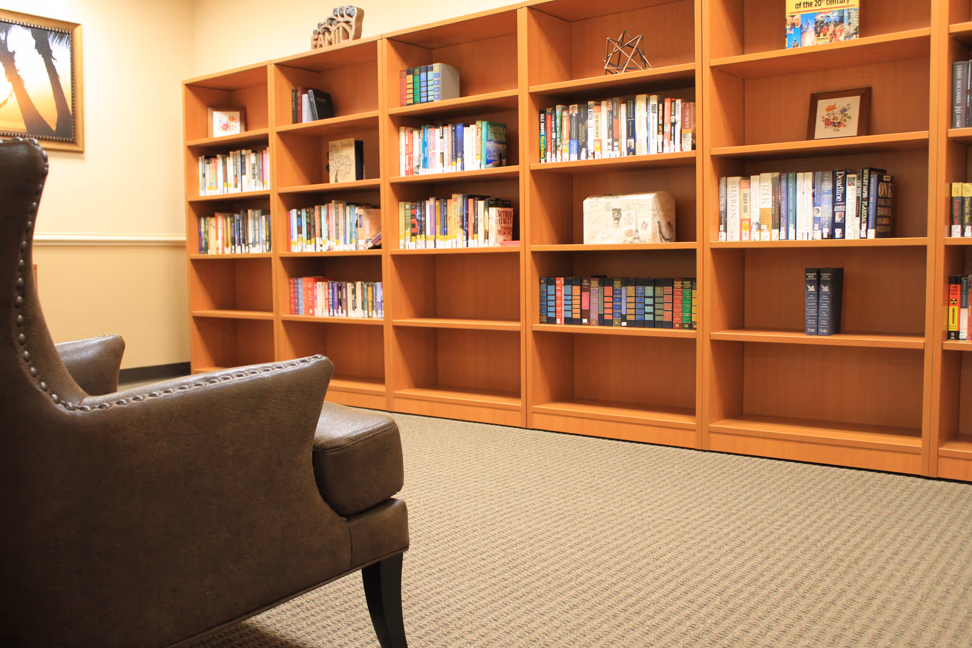 A cozy reading area with a brown leather armchair facing wooden bookshelves filled with books and decorative items in a well-lit room with beige walls and carpeted floor.