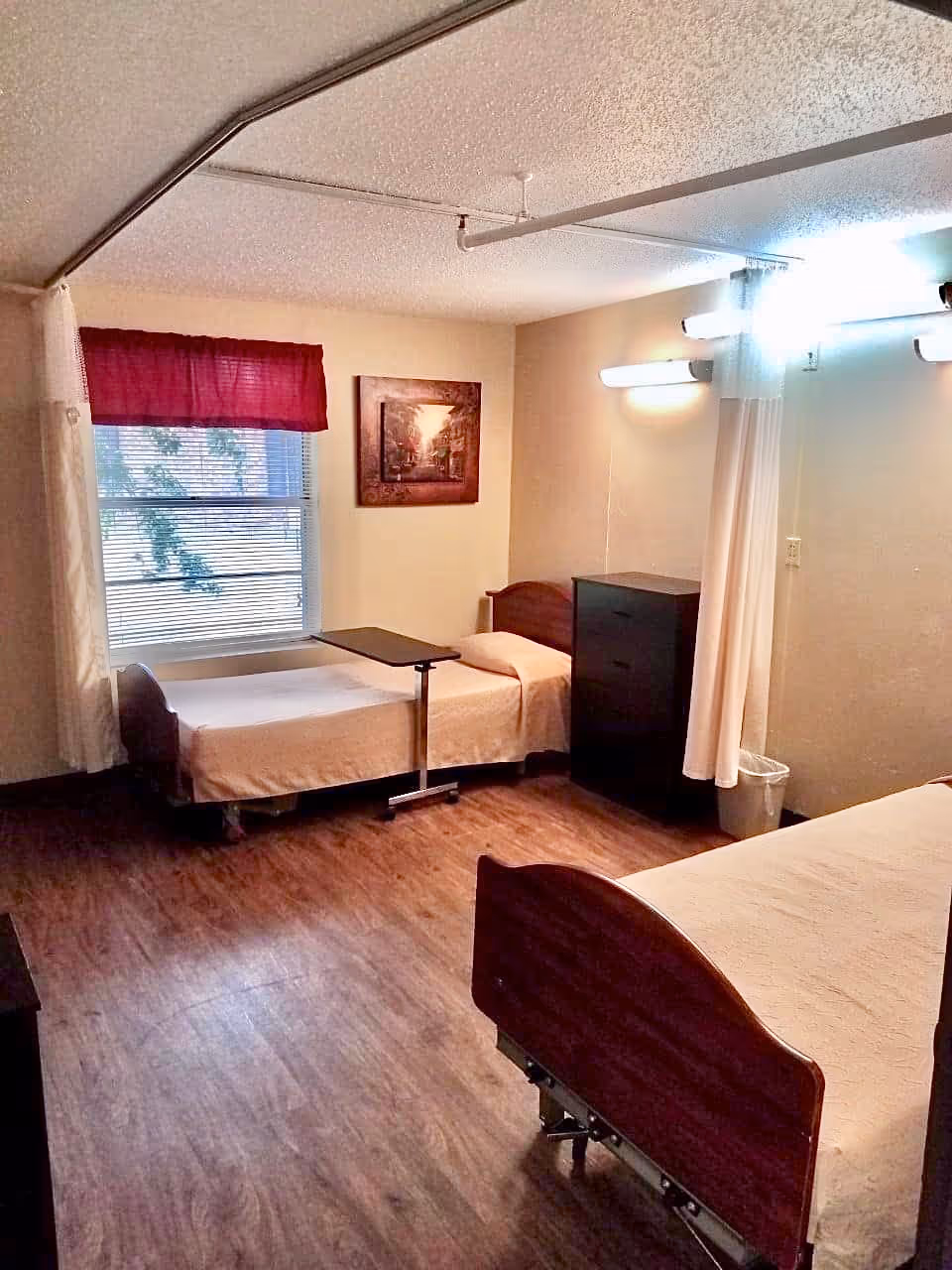 A nursing facility room with two single beds, one near a window with a red valance and blinds, and the other partially visible in the foreground. The room has wood flooring, a small black dresser, a trash can, a wall-mounted light fixture, a curtain divider, and a framed picture on the wall.