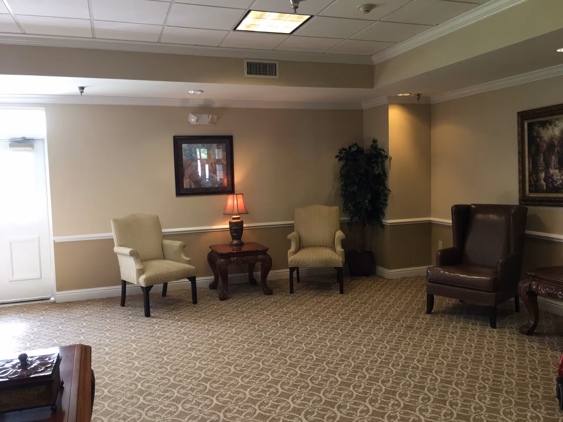 A cozy sitting area with three chairs arranged around two wooden side tables, one with a small lamp. The room has beige walls with white trim, patterned carpet, a potted plant in the corner, and framed artwork on the walls. A door is visible on the left side.