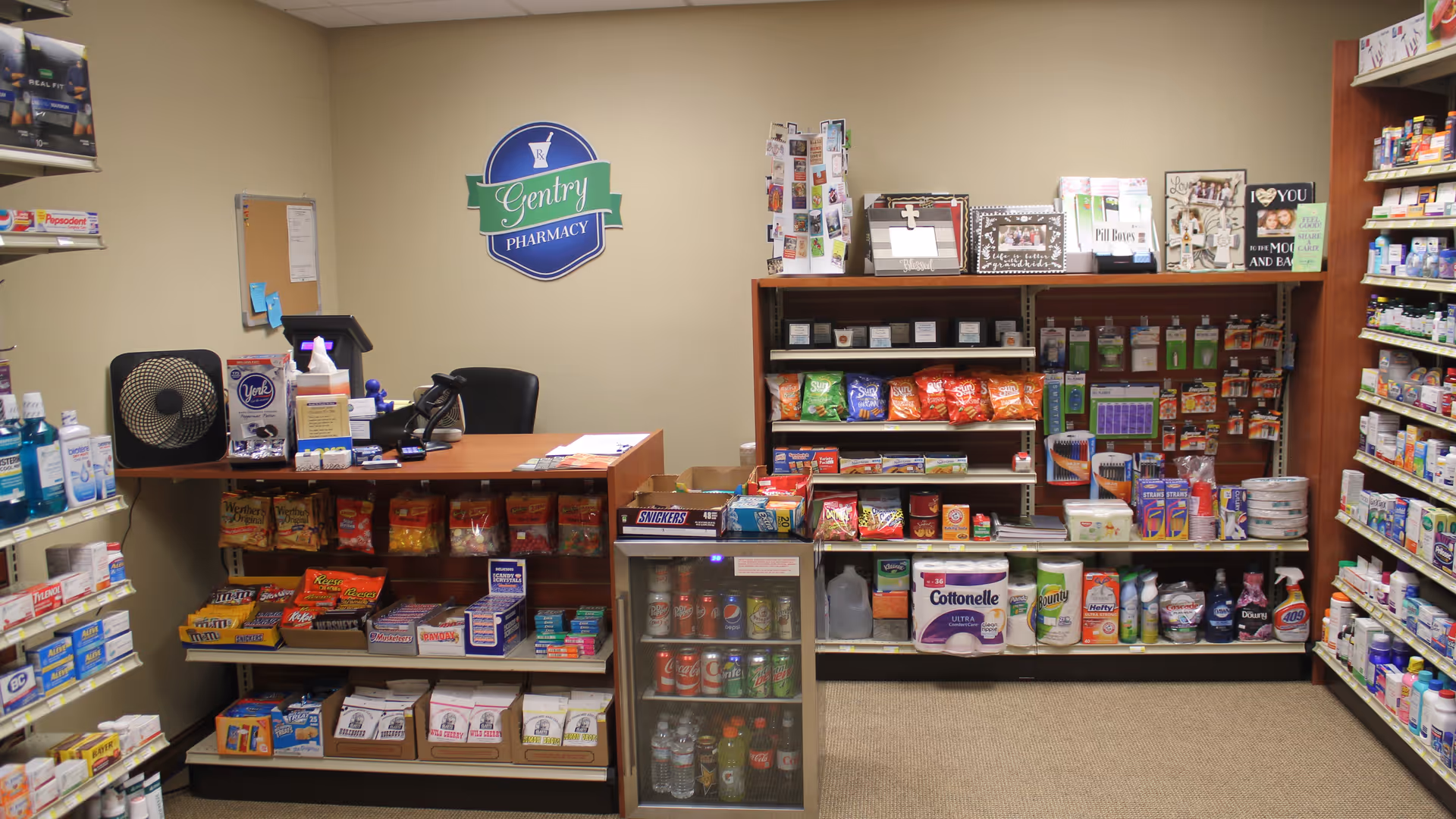 Interior view of a small pharmacy area with shelves stocked with snacks, beverages, toiletries, and over-the-counter medications. A counter with a chair, a fan, and a cash register is visible. A sign on the wall reads 'Gentry Pharmacy'.