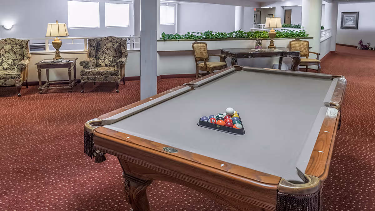 Interior communal lounge with a pool table in the foreground and upholstered chairs, tables, and lamps in the background.