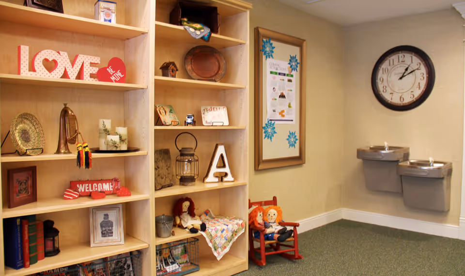 Interior corner of a senior living facility with wooden shelves displaying decorative items such as a 'LOVE' sign, candles, a lantern, and dolls. There is a framed bulletin board with snowflake decorations on the wall and a large round clock showing the time as 12:05. Two wall-mounted water fountains are also visible.