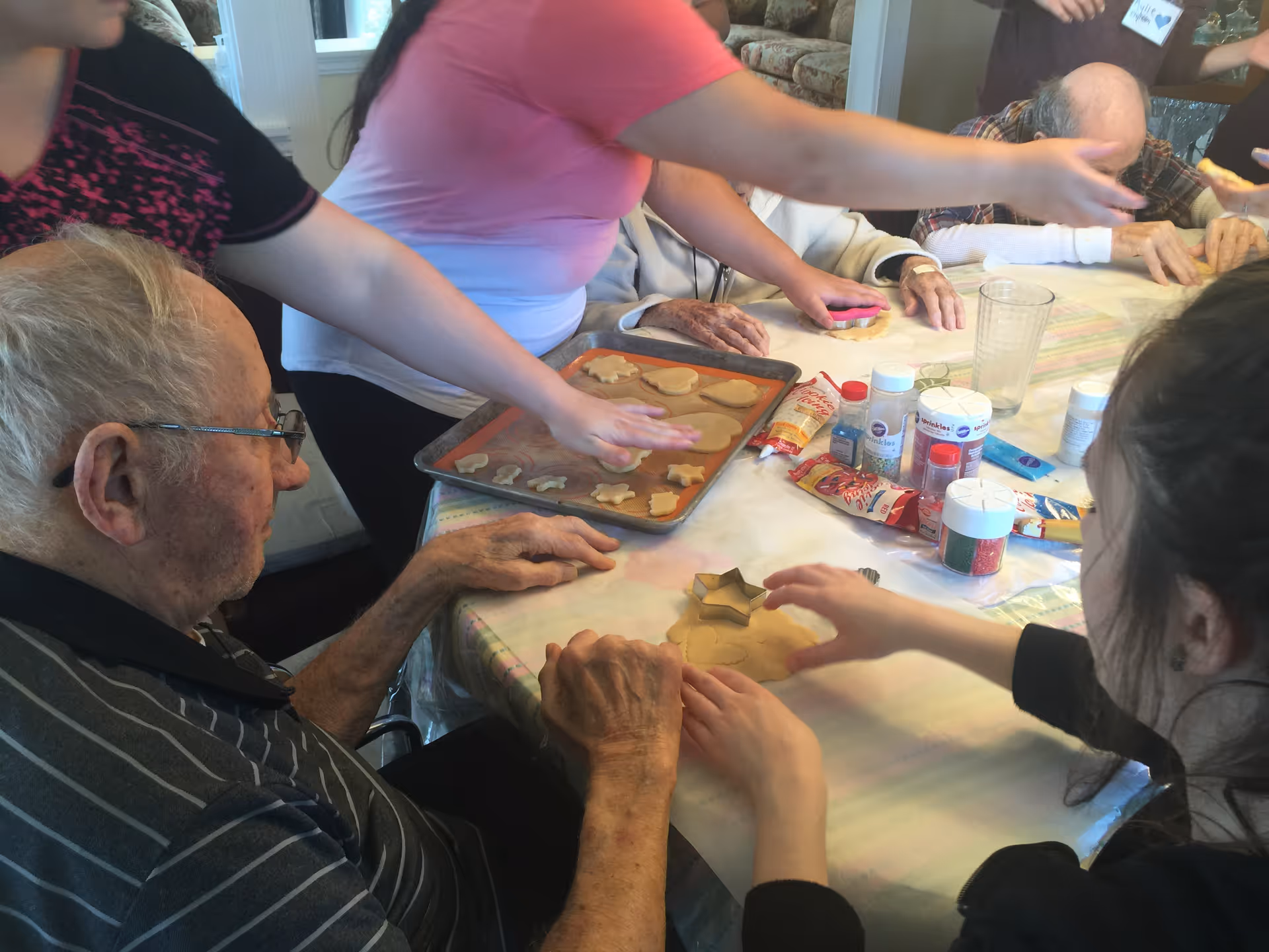 A group of elderly people and caregivers sitting around a table engaged in a cookie decorating activity. Various cookie cutters, sprinkles, and baking supplies are spread out on the table.