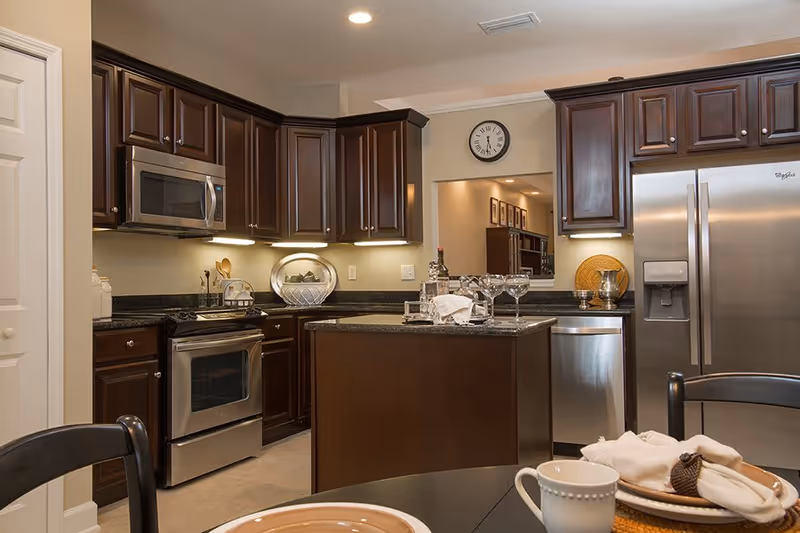 Modern kitchen with dark wooden cabinets, stainless steel appliances including a microwave, oven, dishwasher, and refrigerator. A kitchen island with a tray holding a bottle of wine and four wine glasses is in the center. In the foreground, part of a dining table is visible with a cup, plate, and napkin. A clock is mounted on the wall above a pass-through window to another room.