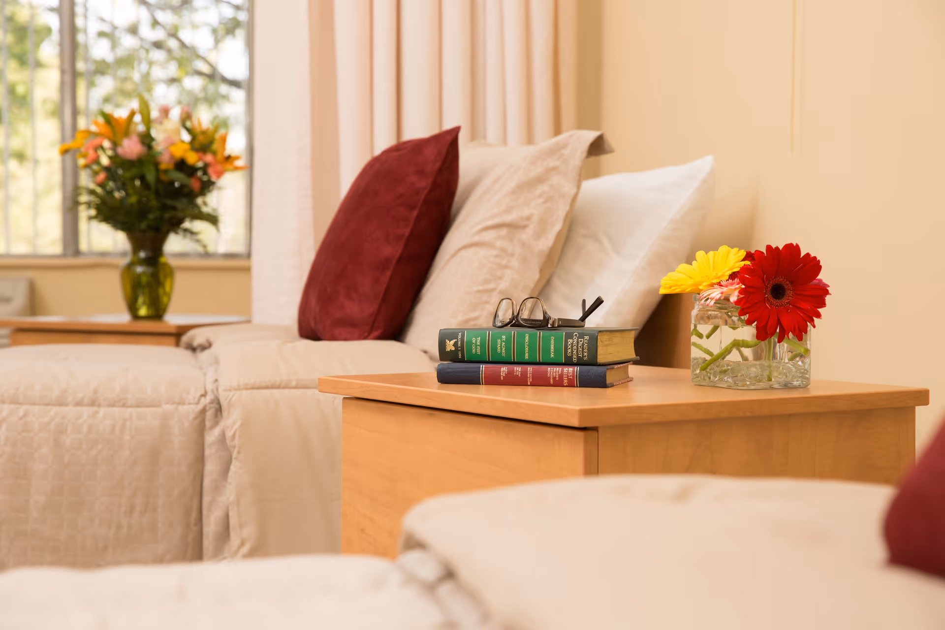 A cozy bedroom corner with a wooden nightstand holding a small glass vase with red and yellow flowers, two stacked books with reading glasses on top, and a beige bed with pillows including a red one. In the background, there is a window with a green vase of flowers on a table.