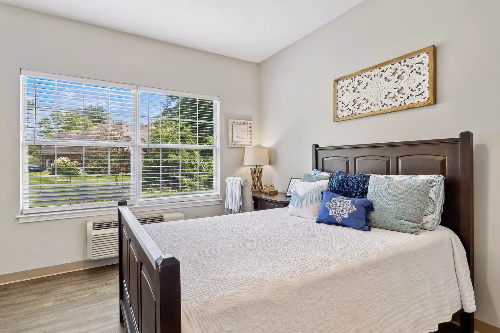 A bright bedroom with a large window showing a green outdoor view. The room features a dark wooden bed frame with a white quilt and several decorative pillows in shades of blue and green. There is a nightstand with a lamp, a small stack of books, and a framed picture. A decorative white and wood wall art piece hangs above the bed.