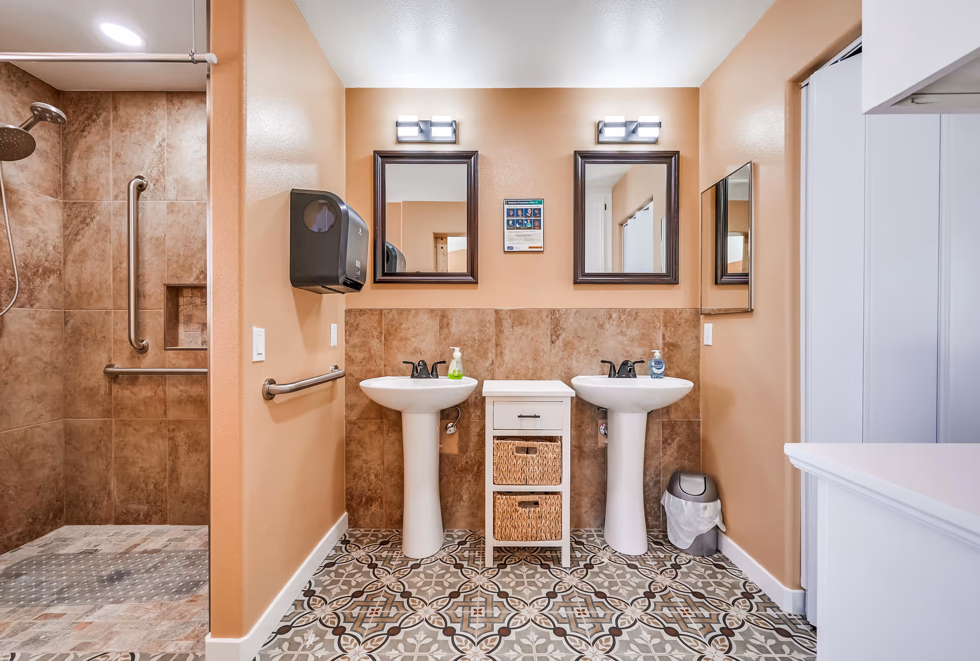 A clean bathroom with two pedestal sinks, each with a mirror and light fixture above. Between the sinks is a small white cabinet with two wicker baskets. To the left is a walk-in shower with tiled walls and a grab bar. The floor has decorative patterned tiles, and the walls are painted a warm beige color. A soap dispenser is mounted on the wall near the left sink, and a trash can is placed near the right sink.