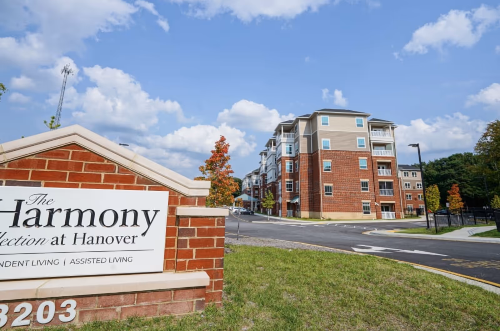Entrance sign for The Harmony Collection at Hanover in front of a multi-story brick senior living building under a blue sky.