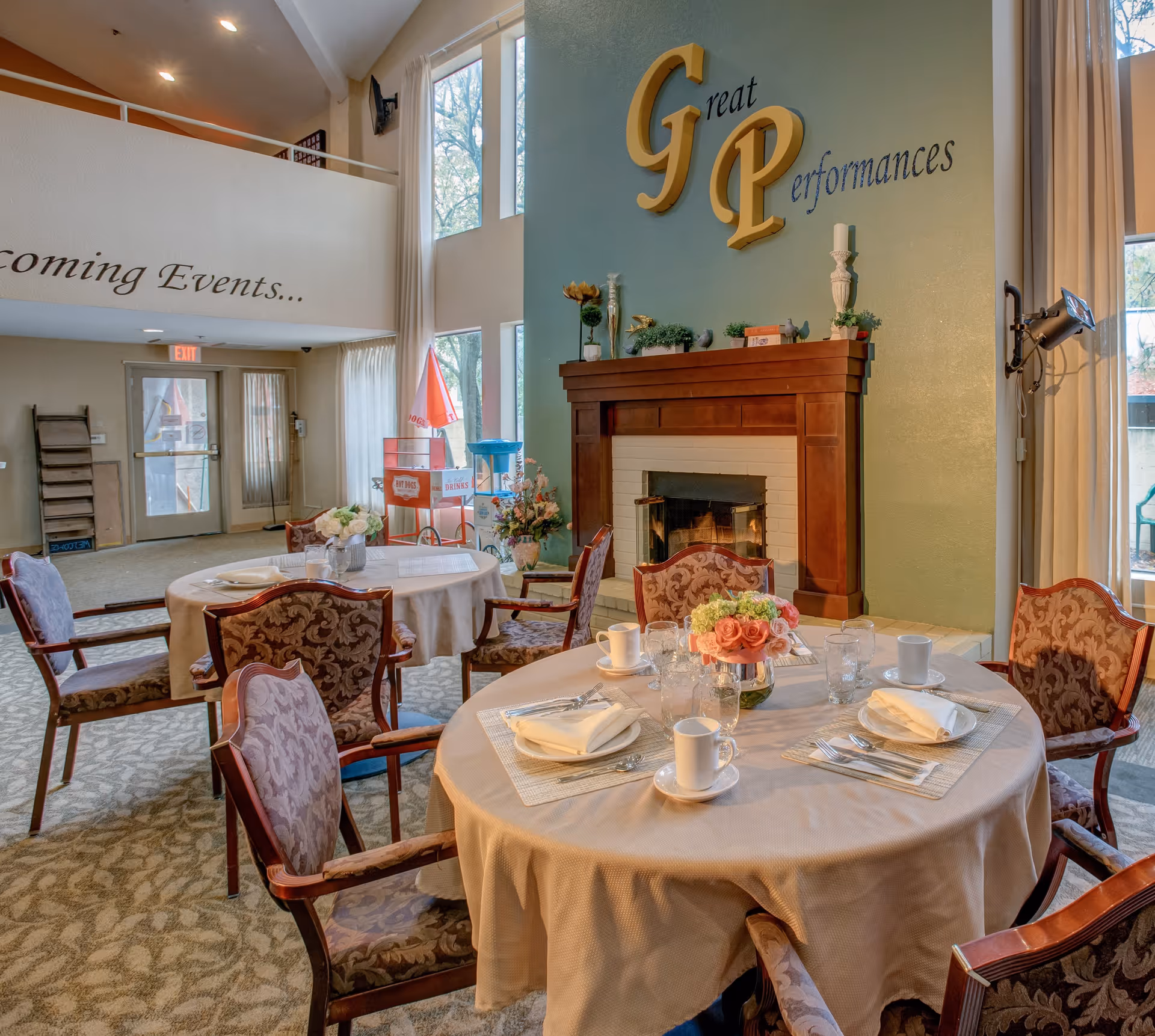 Dining area with round tables set for a meal, upholstered chairs, and a fireplace beneath a wall sign reading 'Great Performances'.