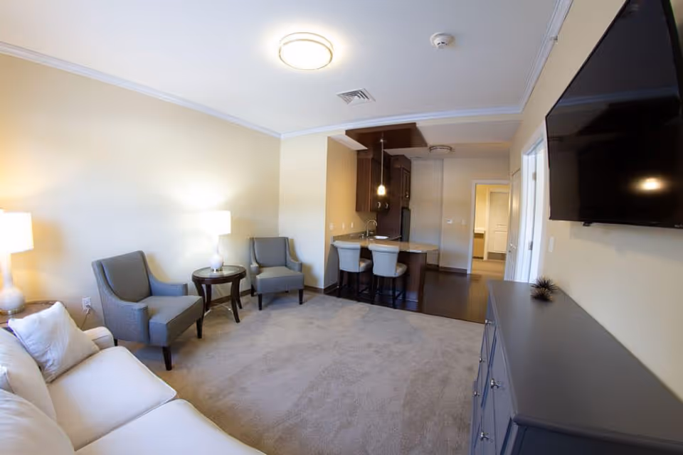 A living room area with a white sofa, two gray armchairs, a round side table with a lamp, and a flat-screen TV mounted on the wall. The room has beige walls and carpeted flooring. In the background, there is a small kitchen area with a breakfast bar and two bar stools.