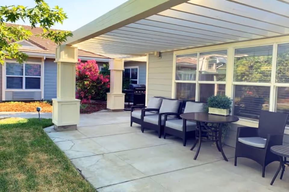 Outdoor patio area at Lassen House Senior Living with a pergola overhead, cushioned chairs, round tables, and a barbecue grill. The patio is adjacent to a building with large windows and surrounded by greenery and flowering bushes.