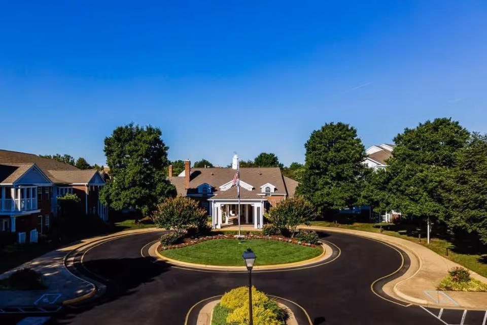 Exterior view of Chancellor's Village senior living facility showing a circular driveway with a flagpole in the center, surrounded by well-maintained landscaping and trees, with residential buildings in the background under a clear blue sky.