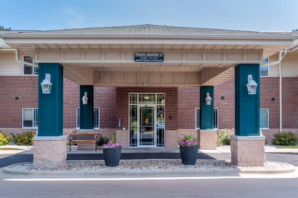 Entrance of Prairie Meadows senior living facility showing a covered drop-off area with four green pillars, two large flower pots with pink flowers, a bench, and a glass door entrance. The building exterior is brick with beige stone accents and has a sign above the entrance reading 'Prairie Meadows III W168 - N11374 Western Avenue'.