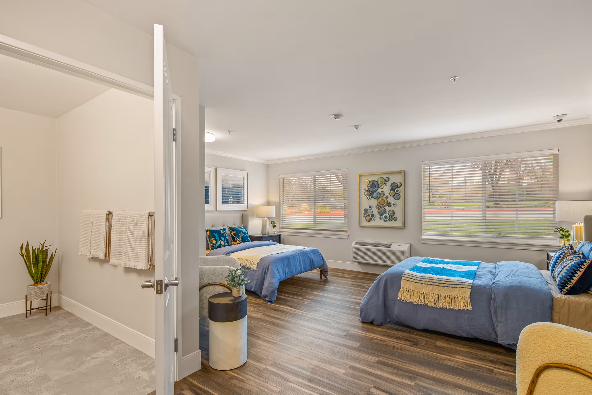 Bright, spacious double bedroom with two made beds, nightstands, seating, artwork, and large windows overlooking a lawn.